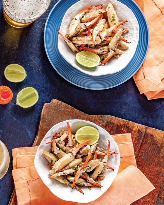 Chile-LimeCrabClaws2 Two bowls of crab meat salad with red pepper strips and herbs, lime wedges, on a dark blue table with orange napkins nearby.
