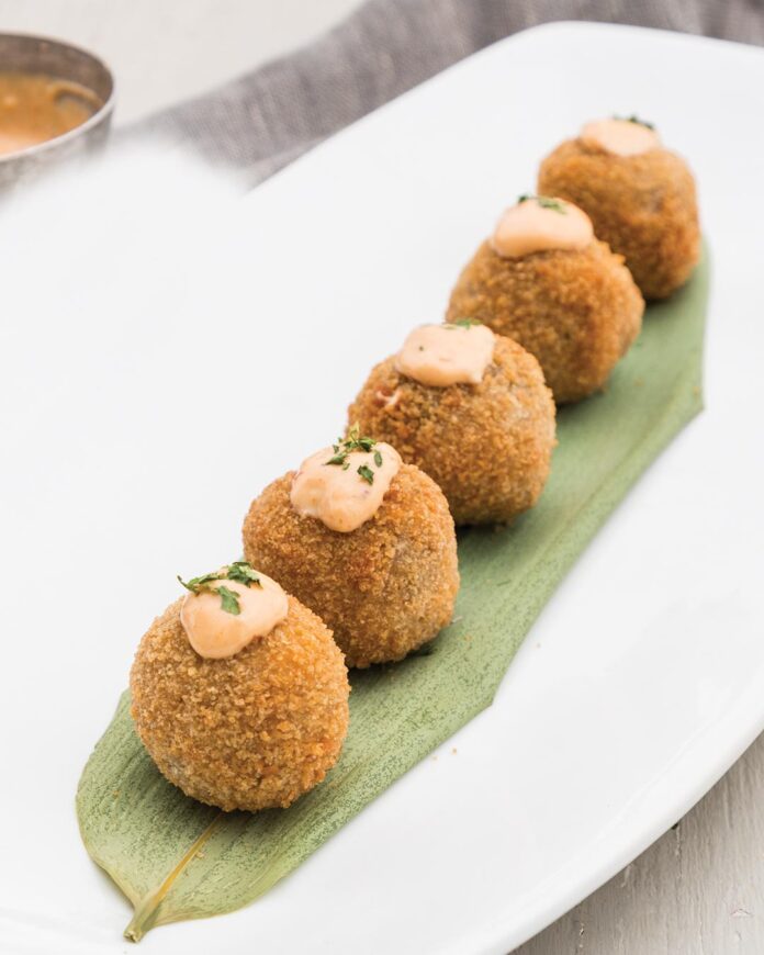 Five breaded croquettes lined on a green leaf with small dollops of creamy orange sauce on top, ready to serve.