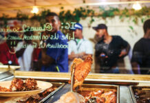 Talking Turkey with Madonna Broussard Gloved hand with tongs lifts glazed chicken from a buffet tray as customers are reflected in the glass behind.