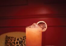 The Dizzie Lizzie Tall glass of iced cocktail with a lime wheel garnish, placed on a wooden table beside halved lime and sliced jalapeños.