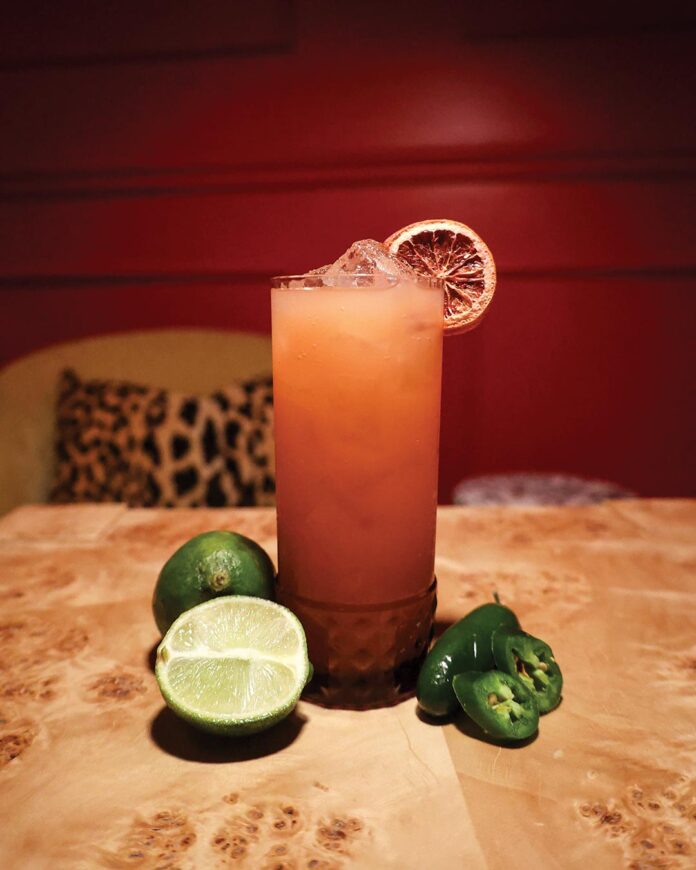 Tall glass of iced cocktail with a lime wheel garnish, placed on a wooden table beside halved lime and sliced jalapeños.
