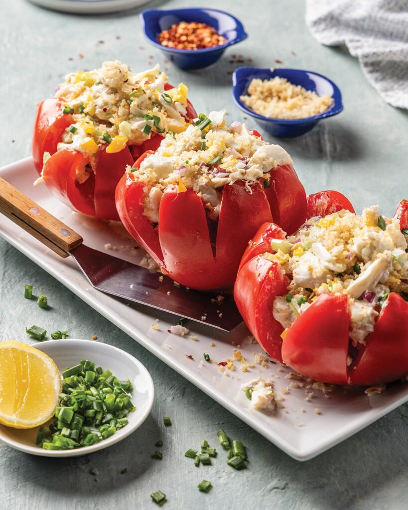 Three hollowed red tomatoes filled with crab salad, egg, and herbs on a white platter, with lemon and chives nearby