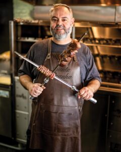 New + Interesting May/June 2026 - Louisiana Cookin' Catch up on all the latest happenings across the Bayou State, with must-try restaurants and so much more. Professional chef in a brown apron grilling skewered meat in a commercial kitchen.