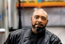 Chef Chat: Lyle Broussard Male chef in a black uniform stands in a commercial kitchen, looking at the camera with a calm expression.