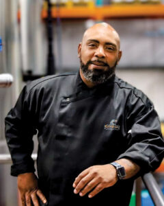Chef Chat: Lyle Broussard - Louisiana Cookin' Chef Lyle Broussard lets us in on the secret to success. Male chef in a black uniform stands in a commercial kitchen, looking at the camera with a calm expression.