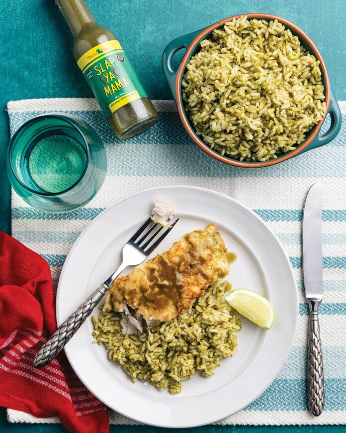 Plate with herb-seasoned rice and a breaded chicken fillet with a lime wedge and fork on a white plate, set on a striped turquoise tablecloth; bottle of Slap Ya Mama pepper sauce and a blue pot of rice nearby, plus a teal glass and red napkin in frame.