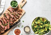 Grilled Marinated Skirt Steak and Smashed Cucumber Salad Sliced medium-rare steak on a wooden board with cilantro, next to a cucumber–avocado salad in a white bowl and small bowls of red pepper flakes and sesame seeds.
