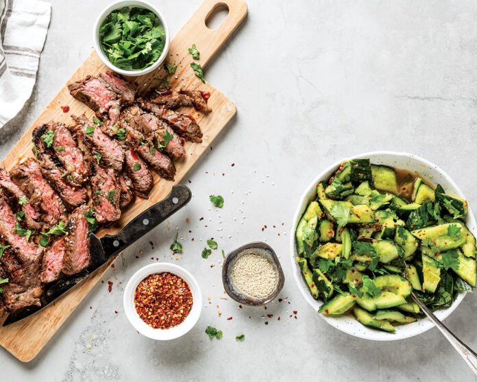 Sliced medium-rare steak on a wooden board with cilantro, next to a cucumber–avocado salad in a white bowl and small bowls of red pepper flakes and sesame seeds.