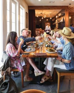Worth the Drive: Small Towns with Big Charm - Louisiana Cookin'  A long wooden table filled with diners in a cozy restaurant, toasting glasses as they share a meal.