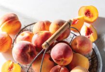 Peach Perfect: 5 Recipes That Make the Most of Summer’s Favorite Fruit Basket of ripe peaches in a wire basket with a wooden-handled fork resting on top; several peaches are halved to reveal orange flesh and red pits nearby.