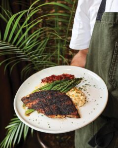 Worth the Drive: Small Towns with Big Charm - Louisiana Cookin'  Chef in white shirt and olive apron presents a plate with herb-crusted fish, asparagus, and risotto.
