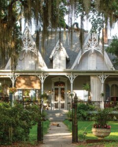 Worth the Drive: Small Towns with Big Charm - Louisiana Cookin'  Victorian-style house with intricate white trim and a wraparound porch, Spanish moss hanging from trees, a cat lying on the path.