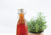Sip and Savor: Sweet Tea-Inspired Recipes for Summer Bottle of red beverage with lemon wedges and rosemary sprig on a light blue tray, with a mason jar of lemons nearby and a potted rosemary plant in the background.