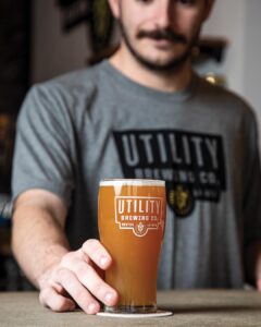 Worth the Drive: Small Towns with Big Charm - Louisiana Cookin'  A man in a gray shirt extends a hand toward a glass of beer on a coaster, beer labeled with Utility Brewing Co. logo in focus.