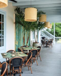 Worth the Drive: Small Towns with Big Charm - Louisiana Cookin'  Outdoor cafe seating on a covered porch with turquoise doors, green tables, and brown wicker chairs under large woven pendant lights.