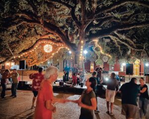 Worth the Drive: Small Towns with Big Charm - Louisiana Cookin'  Outdoor night concert under a large tree with string lights; people dancing while a band plays on a small stage, foregrounded by dancers.