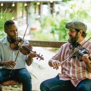 0604TonyManns2777 - Louisiana Cookin' Magic on Lake Martin: Inside Louisiana’s Secret Supper Series - Louisiana Cookin' The guest list is small, the chefs are kept under wraps, and the meal is one to remember. Two men playing violins together outdoors on a wooden bench in a sunlit garden setting