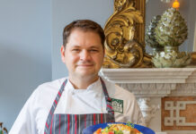 Putting His Heart on the Plate: Inside Chef Jeremy Langlois’ Culinary Journey Male chef in a white uniform and striped gray apron presents a plated dish with colorful vegetables in a formal dining room setting