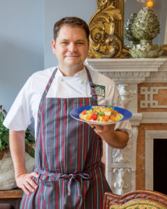 1-JeremyLanglois_179JOH - Louisiana Cookin' Putting His Heart on the Plate: Inside Chef Jeremy Langlois’ Culinary Journey - Louisiana Cookin' Chef Jeremy Langlois reflects on his decades-long culinary journey—from training under legendary John Folse to Houma's House to his latest venture, a self-published cookbook. Male chef in a white uniform and striped gray apron presents a plated dish with colorful vegetables in a formal dining room setting
