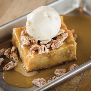 20170316_secondline_0017 - Louisiana Cookin' Making Waves: Louisiana’s Own Culinary Trailblazers - Louisiana Cookin' Louisiana-born chefs making their mark across the South. Square caramel custard cake topped with a scoop of vanilla ice cream and chopped pecans, covered in caramel sauce on a metal tray.