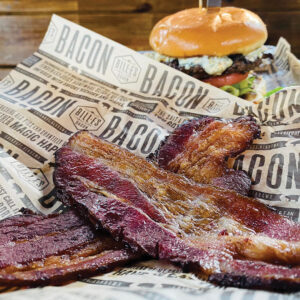 BACONTASTER2021 - Louisiana Cookin' Weekend Away: Press Pause in Fairhope, Alabama - Louisiana Cookin' A gentle weekend escape to a city full of coastal charm. Thick-cut bacon slices in focus on branded parchment, with a burger in the blurred background behind.