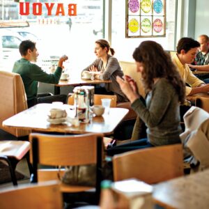 Bayou-Bakery-Interior_Guests1_ScottSuchman - Louisiana Cookin' Making Waves: Louisiana’s Own Culinary Trailblazers - Louisiana Cookin' Louisiana-born chefs making their mark across the South. Two people sit at a wooden table in a bright cafe, chatting over coffee cups while others work or relax nearby.