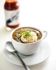 Bayou-Bakery_Gumbo_Rey-Lopez - Louisiana Cookin' Making Waves: Louisiana’s Own Culinary Trailblazers - Louisiana Cookin' Louisiana-born chefs making their mark across the South. Brown broth soup in a white cup topped with a mound of white rice and green onion slices on a white saucer with a spoon nearby.