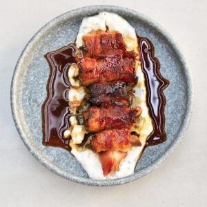 Cajun-Duck-Poppers - Louisiana Cookin' Making Waves: Louisiana’s Own Culinary Trailblazers - Louisiana Cookin' Louisiana-born chefs making their mark across the South. Plate with bacon-wrapped meat skewers on a creamy white sauce, drizzled with dark glaze and a light brown sauce.