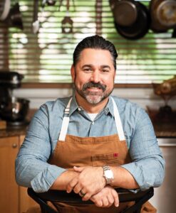 Chef-David-Guas - Louisiana Cookin' Making Waves: Louisiana’s Own Culinary Trailblazers - Louisiana Cookin' Louisiana-born chefs making their mark across the South. Smiling man in a blue denim shirt and brown apron sits with arms crossed in a kitchen behind him.