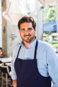 Chef-Drake-Leonards - Louisiana Cookin' Making Waves: Louisiana’s Own Culinary Trailblazers - Louisiana Cookin' Louisiana-born chefs making their mark across the South. Smiling man with a beard wearing a blue checkered shirt and navy apron in a bright cafe setting