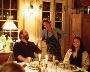 Copy-of-NinaSS2842 - Louisiana Cookin' Magic on Lake Martin: Inside Louisiana’s Secret Supper Series - Louisiana Cookin' The guest list is small, the chefs are kept under wraps, and the meal is one to remember. Three friends at a warmly lit dinner table; a bearded man gestures as a woman in a striped apron laughs beside him.