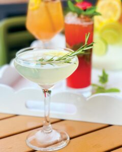 Garden-Party_Neutral-Ground_Simone-Rathle - Louisiana Cookin' Making Waves: Louisiana’s Own Culinary Trailblazers - Louisiana Cookin' Louisiana-born chefs making their mark across the South. Cocktail in a coupe glass garnished with a rosemary sprig and lemon twist on a wooden table, with colorful drinks blurred in the background.