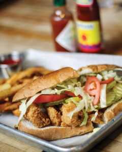 IMG_8660 - Louisiana Cookin' Making Waves: Louisiana’s Own Culinary Trailblazers - Louisiana Cookin' Louisiana-born chefs making their mark across the South. Crispy fried chicken sandwich with lettuce and tomato on a crusty bun, on a metal tray.