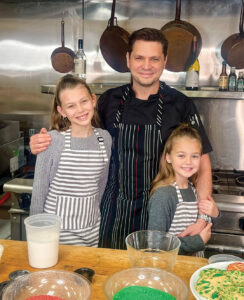 IMG_9709 - Louisiana Cookin' Putting His Heart on the Plate: Inside Chef Jeremy Langlois’ Culinary Journey - Louisiana Cookin' Chef Jeremy Langlois reflects on his decades-long culinary journey—from training under legendary John Folse to Houma's House to his latest venture, a self-published cookbook. Chef with two young girls in striped aprons posing in a stainless-steel kitchen, smiling at the camera.