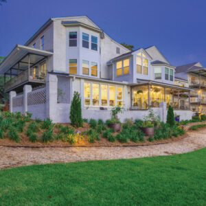 JubileeSuites118-SUB - Louisiana Cookin' Weekend Away: Press Pause in Fairhope, Alabama - Louisiana Cookin' A gentle weekend escape to a city full of coastal charm. Modern two‑story house with large windows and a wraparound porch, illuminated from inside. Lush landscaping and a gravel path in the foreground.
