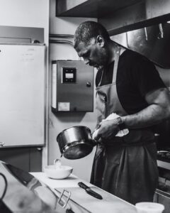 Kenyatta-Heor-Image-2 - Louisiana Cookin' Making Waves: Louisiana’s Own Culinary Trailblazers - Louisiana Cookin' Louisiana-born chefs making their mark across the South. Black-and-white image of a chef in an apron pouring liquid from a pot into a bowl on a kitchen counter, concentrating on the task
