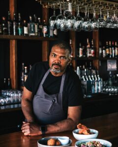 Kenyatta-Hero-Image-1 - Louisiana Cookin' Making Waves: Louisiana’s Own Culinary Trailblazers - Louisiana Cookin' Louisiana-born chefs making their mark across the South. Male bartender or chef in a black shirt and gray apron leans on a wooden bar, with bowls of fried foods in the foreground and bottles on the shelves behind him.