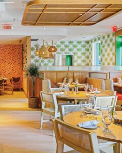 Main-Dining-Cabana-Room-and-Entry_NeutralGround-1 - Louisiana Cookin' Making Waves: Louisiana’s Own Culinary Trailblazers - Louisiana Cookin' Louisiana-born chefs making their mark across the South. Bright modern restaurant dining area with round wooden tables, glassware, and woven pendant lights.