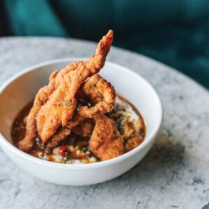 NG-Catfish--Grits - Louisiana Cookin' Making Waves: Louisiana’s Own Culinary Trailblazers - Louisiana Cookin' Louisiana-born chefs making their mark across the South. Crispy fried chicken tenders stacked in a white bowl over a chunky tomato-based sauce with herbs.