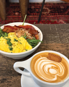 Provision1 - Louisiana Cookin' Weekend Away: Press Pause in Fairhope, Alabama - Louisiana Cookin' A gentle weekend escape to a city full of coastal charm. Bowl of scrambled eggs with greens and crispy bacon, garnished with herbs, next to a latte with swirled foam.