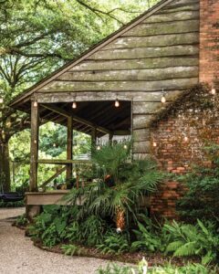 SSSDrakeLeonards_5931-1 - Louisiana Cookin' Magic on Lake Martin: Inside Louisiana’s Secret Supper Series - Louisiana Cookin' The guest list is small, the chefs are kept under wraps, and the meal is one to remember. Rustic wooden cabin porch with string lights, lush greenery, and a brick wall on the right; a gravel path nearby.