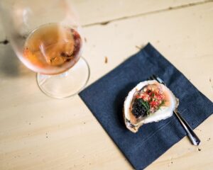 SSSDrakeLeonards_5949 - Louisiana Cookin' Magic on Lake Martin: Inside Louisiana’s Secret Supper Series - Louisiana Cookin' The guest list is small, the chefs are kept under wraps, and the meal is one to remember. Oyster on a half shell topped with herbs and red accents, placed on a dark napkin beside a glass of pink wine on a light wooden table.