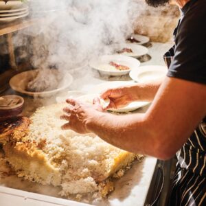 SS_BrushlandSohailZandi3727 - Louisiana Cookin' Magic on Lake Martin: Inside Louisiana’s Secret Supper Series - Louisiana Cookin' The guest list is small, the chefs are kept under wraps, and the meal is one to remember. Chef grating or shredding coconut on a counter as steam rises, with bowls and plates in the background.
