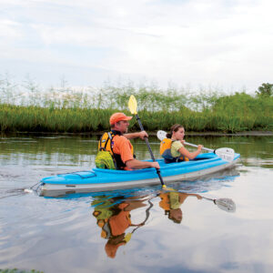 Spanish-Fort-Kayaking-in-Delta - Louisiana Cookin' Weekend Away: Press Pause in Fairhope, Alabama - Louisiana Cookin' A gentle weekend escape to a city full of coastal charm. Two people in an orange life jacket and a yellow-green life jacket paddle a blue tandem kayak on a calm river, with reeds along the shore and their reflections in the water.