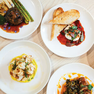 Top-Down-Spring-Menu-108-2-1 - Louisiana Cookin' Weekend Away: Press Pause in Fairhope, Alabama - Louisiana Cookin' A gentle weekend escape to a city full of coastal charm. Assorted plated dishes on a light wood table: braised beef with asparagus and potatoes, served with red sauce on the left plate; bruschetta-style plate with burrata or goat cheese over tomato sauce and greens; yellow pasta with shrimp on another plate; meat with greens in an orange sauce on the last plate.