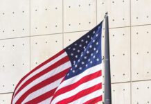 America 250: A Guide to Louisiana’s Semiquincentennial Celebrations American flag waving on a flagpole in front of a light beige wall with small circular fixtures.