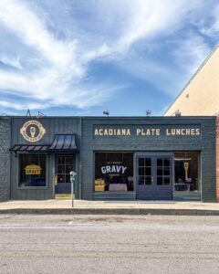 web-Gravy2CreditTrevorJohnsonMaraisStudios - Louisiana Cookin' New + Interesting: July/August 2026 - Louisiana Cookin' Catch up on all the latest happenings across the Bayou state, with must-try restaurants and so much more. Blue-gray brick diner facade with 'ACADIANA PLATE LUNCHES' sign; left window shows circular 'Gravy Lafayette' logo, center window displays 'GRAVY' sign.