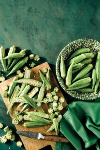 Okra State of Mind: 5 Recipes That Make the Most of Summer Okra - Louisiana Cookin' This fuzzy green pod thrives in the thick of the summer. Fresh okra pods on a wooden cutting board and in a patterned bowl against a green backdrop, with a knife nearby.