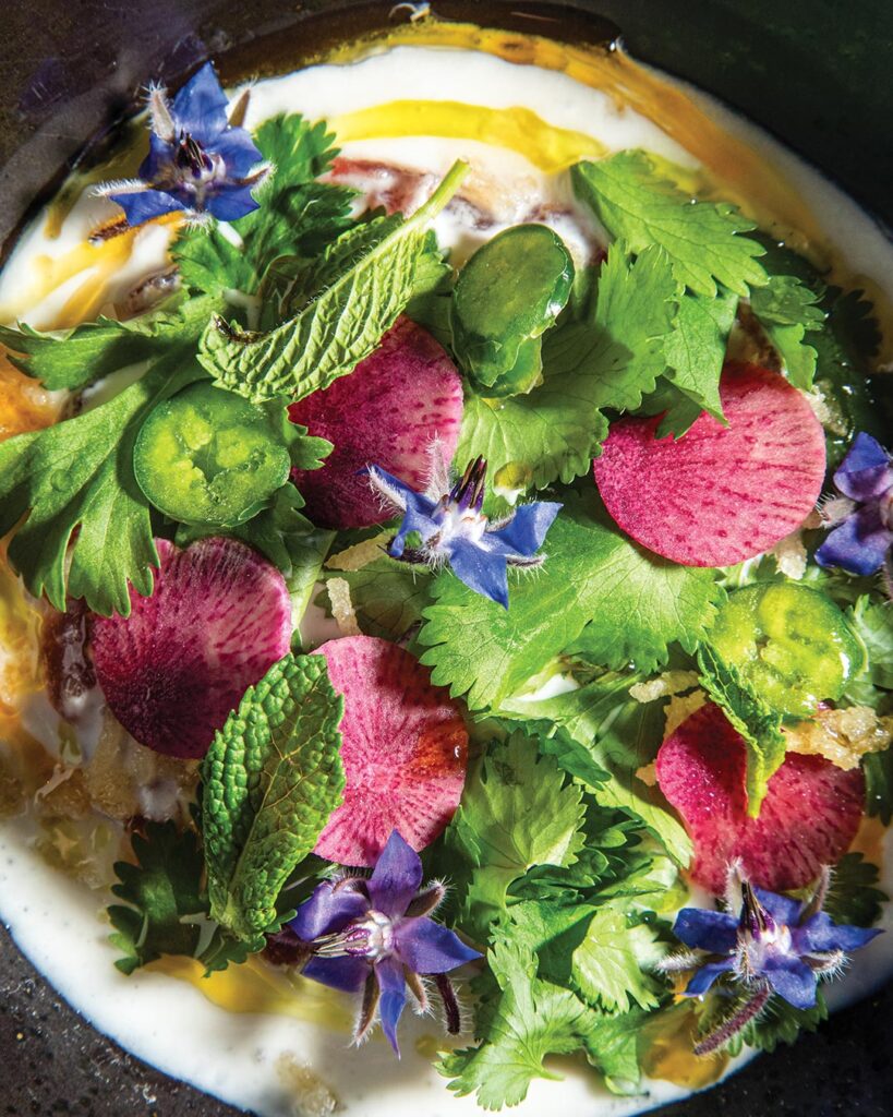 Colorful herb salad with pink radish slices, green herbs, and blue edible flowers atop a creamy base in a bowl
