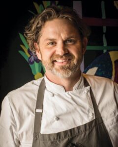 webKelly-English - Louisiana Cookin' Making Waves: Louisiana’s Own Culinary Trailblazers - Louisiana Cookin' Louisiana-born chefs making their mark across the South. Smiling male chef in a white chef coat and dark apron, posing against a colorful mural background.